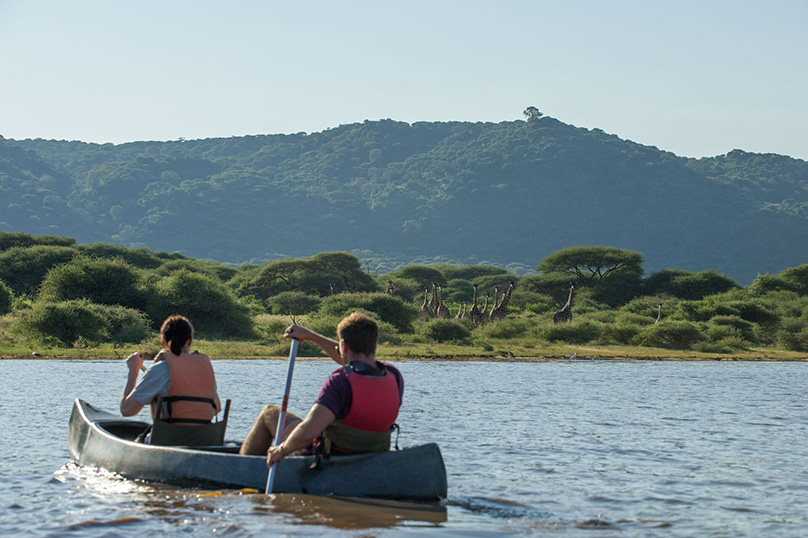 Kanufahrer auf Manyara-See mit Giraffen und Manyara Escarpment im Hintergrund